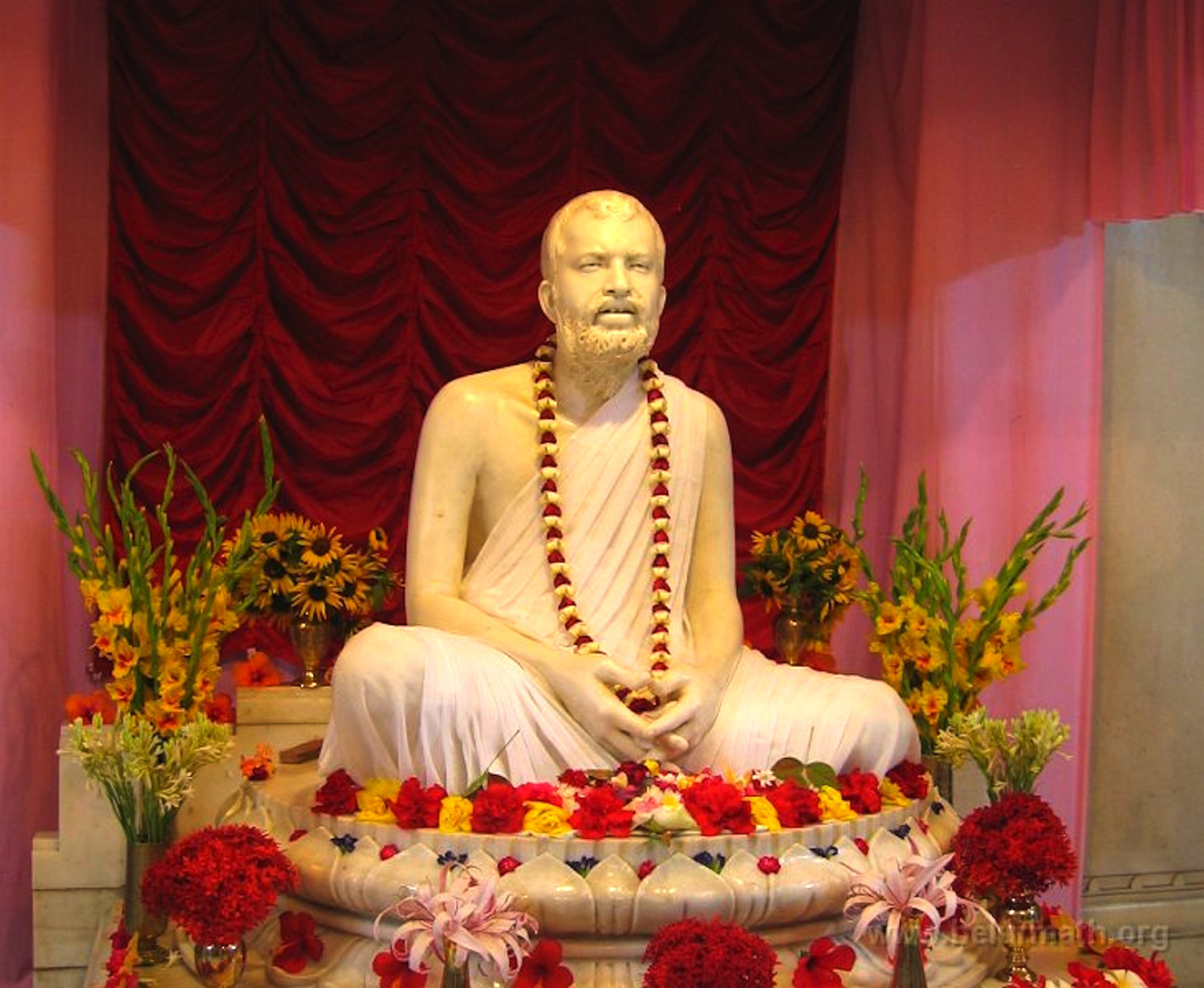 Scupture of Sri Ramakrishna seated with a flower necklasurrounded by flowers.